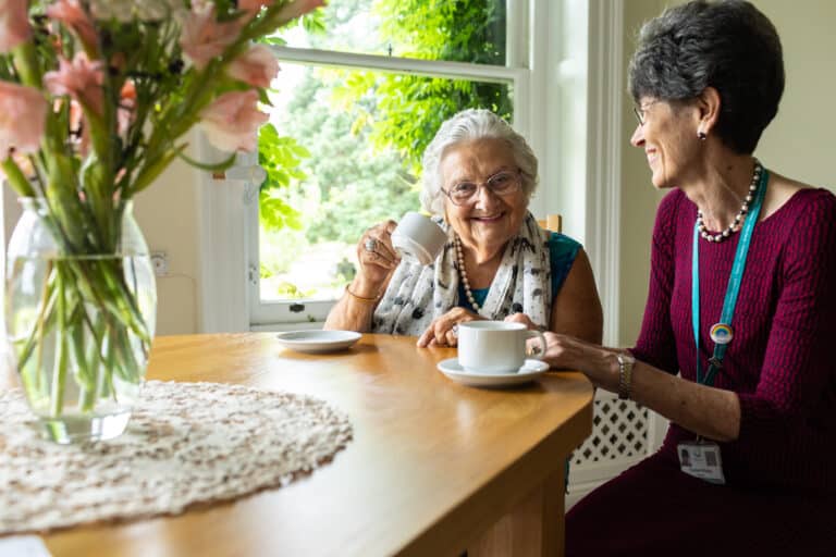 Elderly resident sat at a table adorned by fresh flowers having a cup of tea with one of the Directors at Birtley House Nursing Home, giving a feeling of extended family