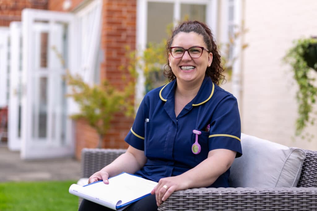 A smiling nurse sitting on a garden sofa with Birtley House Care Home in the background